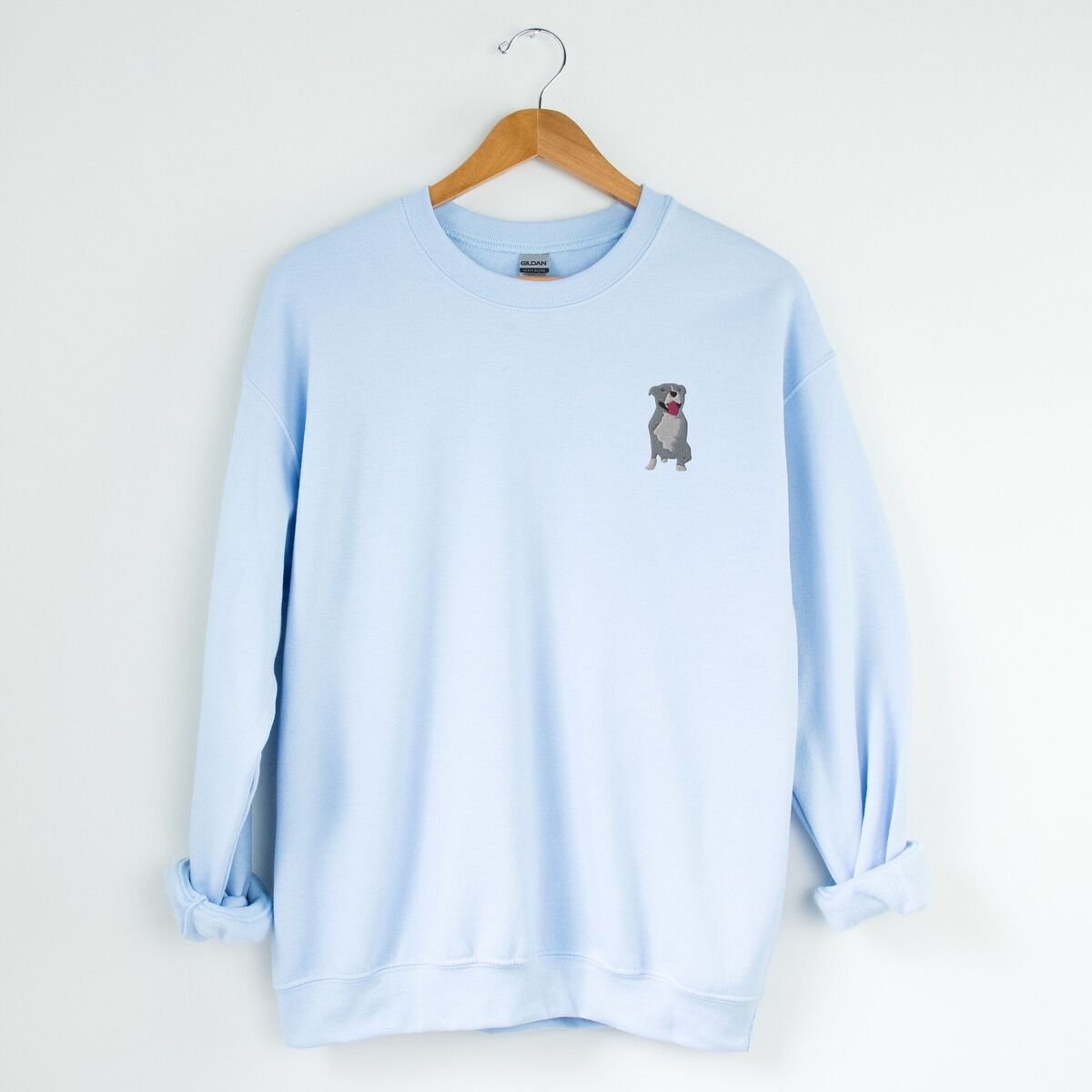 Blue Pitbull Sweatshirt - Light Blue Light Blue crewneck sweatshirt on a hanger, embroidered with a small blue-grey pit bull sitting with its pink tongue out on the left chest