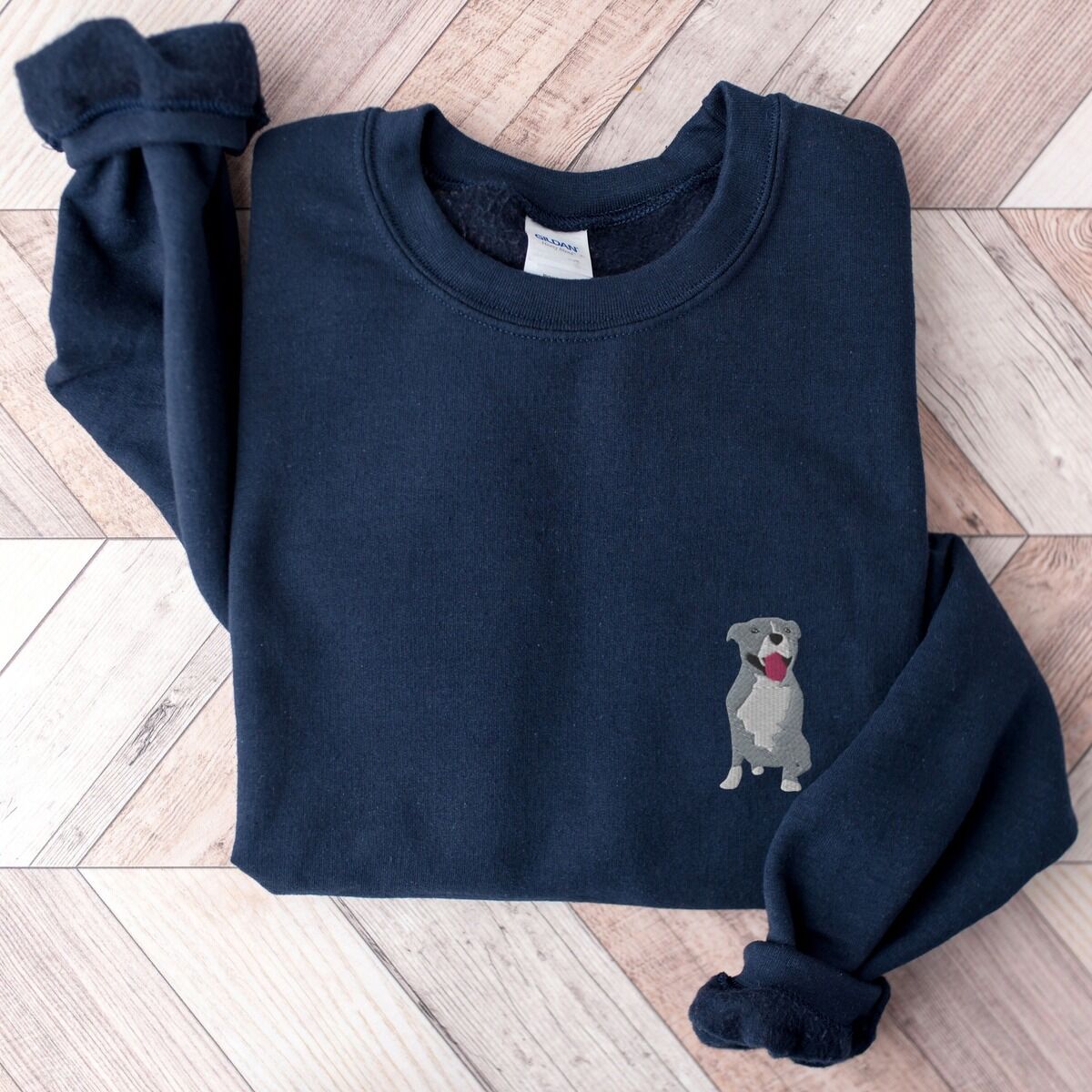 Blue Pitbull Sweatshirt – Navy Navy crewneck sweatshirt laid flat on a wood floor, embroidered with a small blue-grey pit bull sitting with its pink tongue out on the left chest