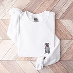 White crewneck sweatshirt laid flat on a wood floor, embroidered with a small blue-grey pit bull sitting with its pink tongue out on the left chest
