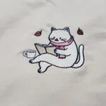 Close-up of the embroidered bookworm cat in a pink scarf reading an open book with a coffee cup and two autumn leaves