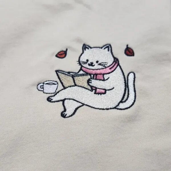Close-up of the embroidered bookworm cat in a pink scarf reading an open book with a coffee cup and two autumn leaves