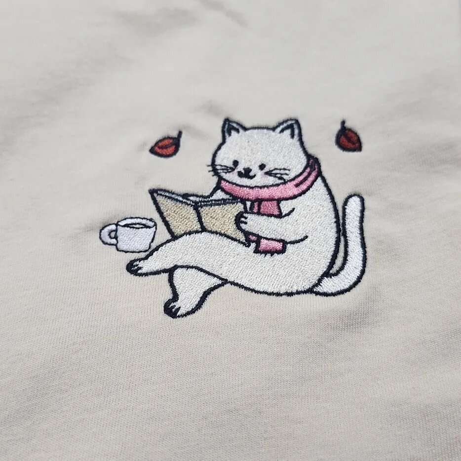 Bookworm Cat Embroidery – Close-up Close-up of the embroidered bookworm cat in a pink scarf reading an open book with a coffee cup and two autumn leaves
