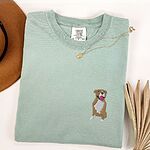Bay T-shirt laid flat with a gold necklace and dried palm leaves, embroidered with a small fawn and white pit bull sitting with its pink tongue out on the left chest