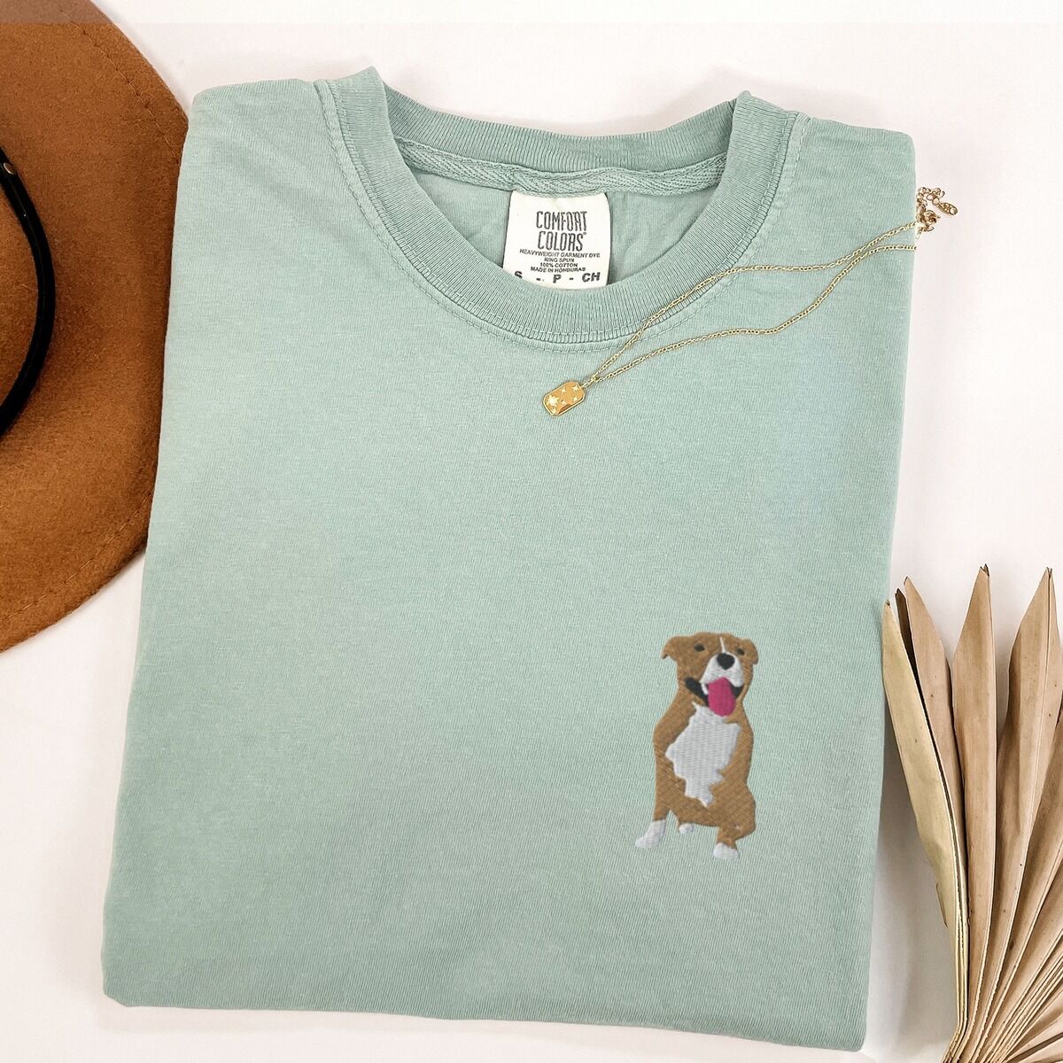 Brown and White Pitbull Shirt – Bay Bay T-shirt laid flat with a gold necklace and dried palm leaves, embroidered with a small fawn and white pit bull sitting with its pink tongue out on the left chest