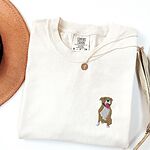 Ivory T-shirt laid flat with a straw hat and a gold necklace, embroidered with a small fawn and white pit bull sitting with its pink tongue out on the left chest