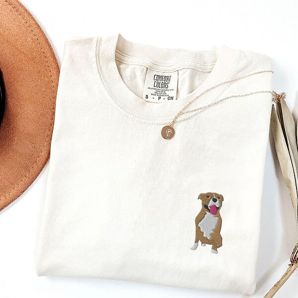 Ivory T-shirt laid flat with a straw hat and a gold necklace, embroidered with a small fawn and white pit bull sitting with its pink tongue out on the left chest
