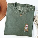 Moss T-shirt laid flat with a gold necklace and dried palm leaves, embroidered with a small fawn and white pit bull sitting with its pink tongue out on the left chest