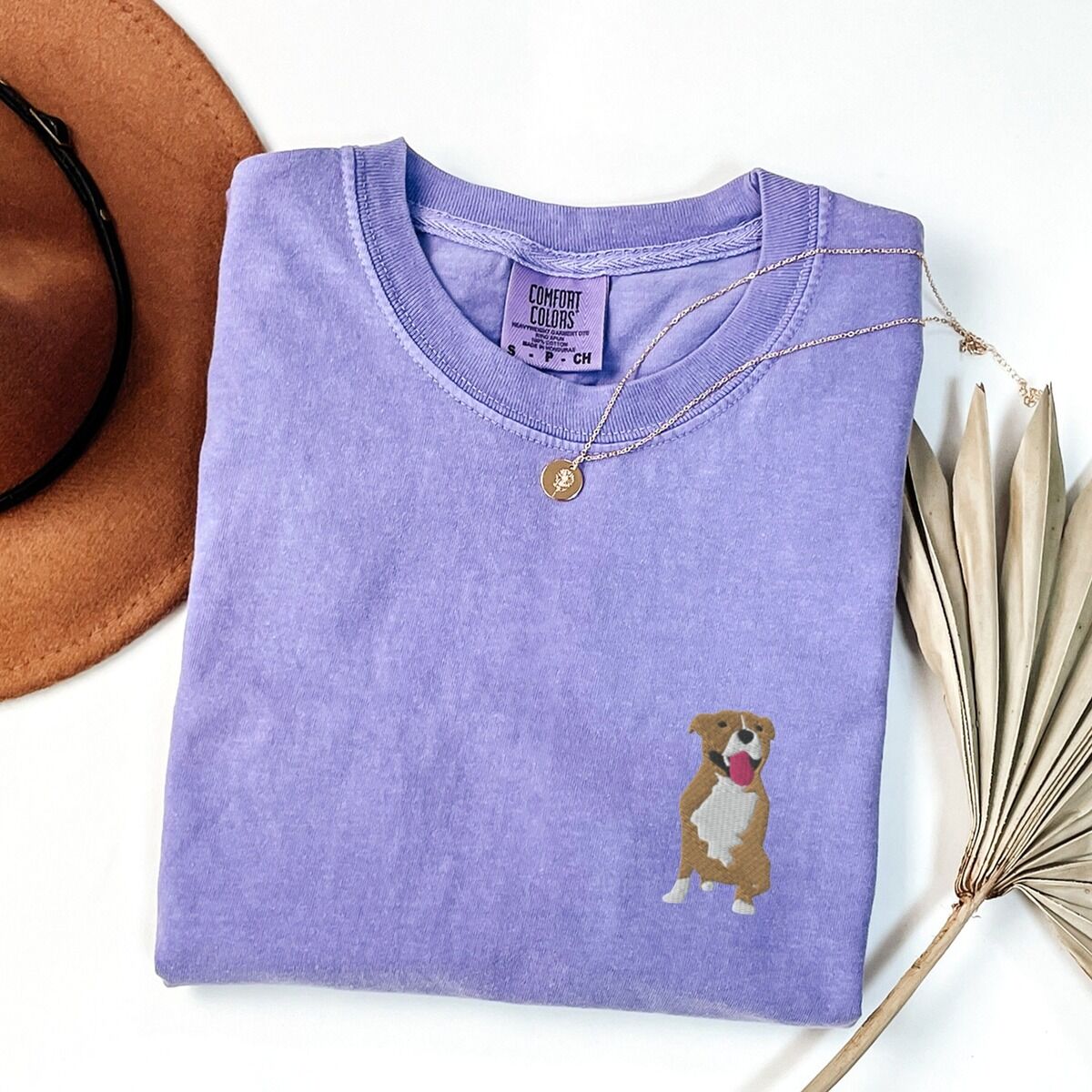 Brown and White Pitbull Shirt – Violet Violet T-shirt laid flat with a straw hat and a gold necklace, embroidered with a small fawn and white pit bull sitting with its pink tongue out on the left chest