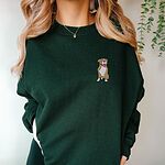 Woman in a Forest Green crewneck sweatshirt embroidered with a small fawn and white pit bull sitting with its pink tongue out on the left chest
