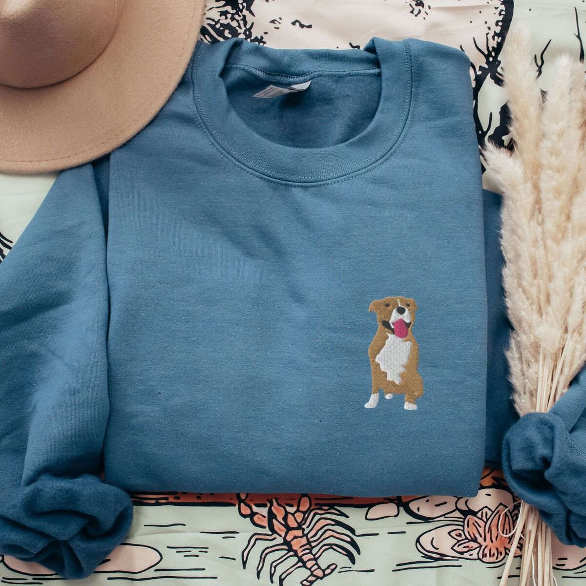 Brown and White Pitbull Sweatshirt – Indigo Blue Indigo Blue crewneck sweatshirt laid flat with a straw hat and dried pampas, embroidered with a small fawn and white pit bull sitting with its pink tongue out on the left chest