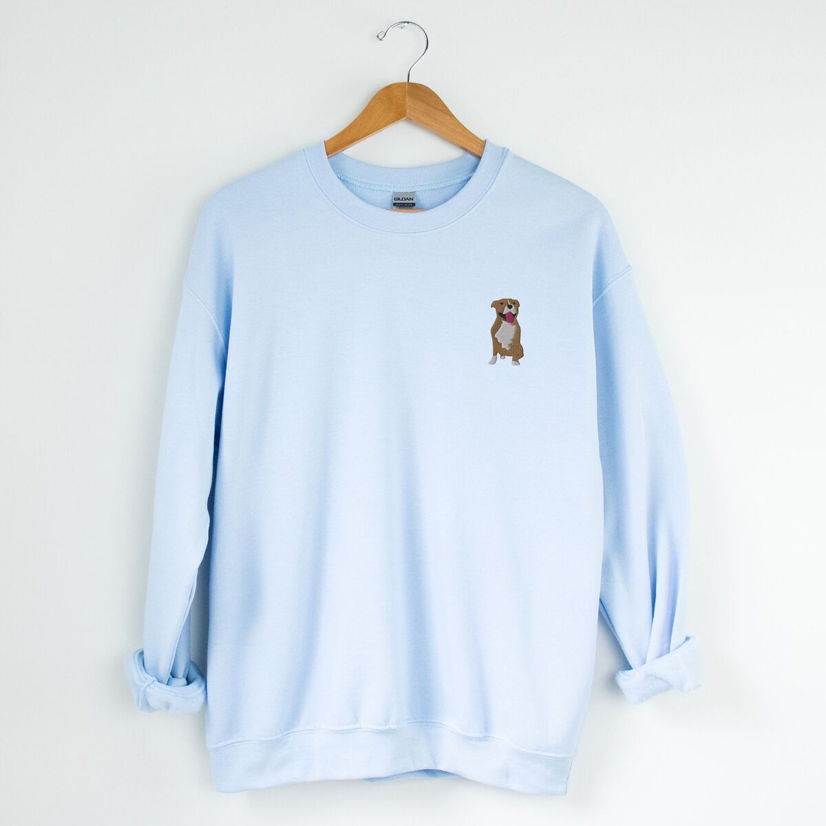 Brown and White Pitbull Sweatshirt - Light Blue Light Blue crewneck sweatshirt on a hanger, embroidered with a small fawn and white pit bull sitting with its pink tongue out on the left chest