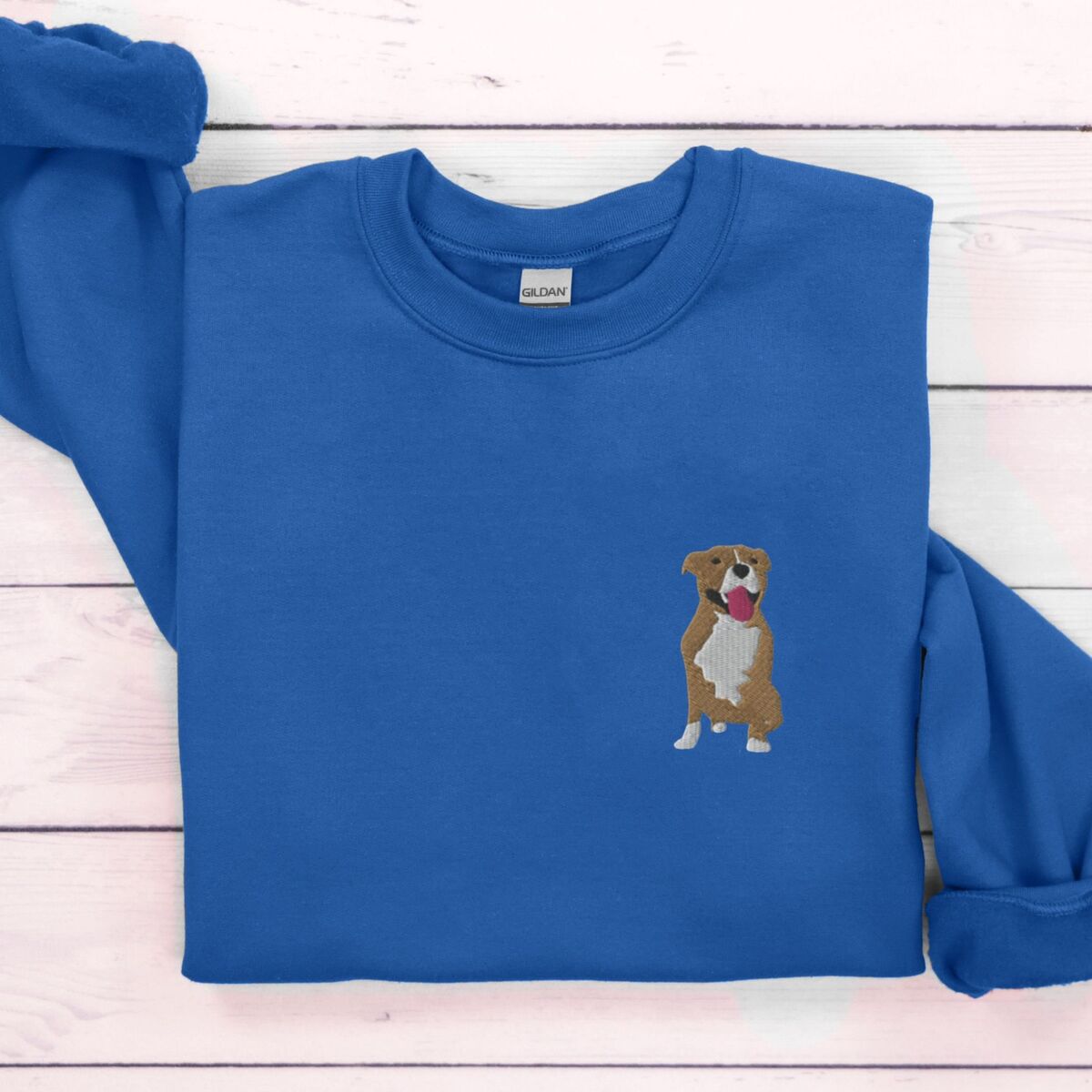 Brown and White Pitbull Sweatshirt - Royal Royal crewneck sweatshirt laid flat on white wood, embroidered with a small fawn and white pit bull sitting with its pink tongue out on the left chest