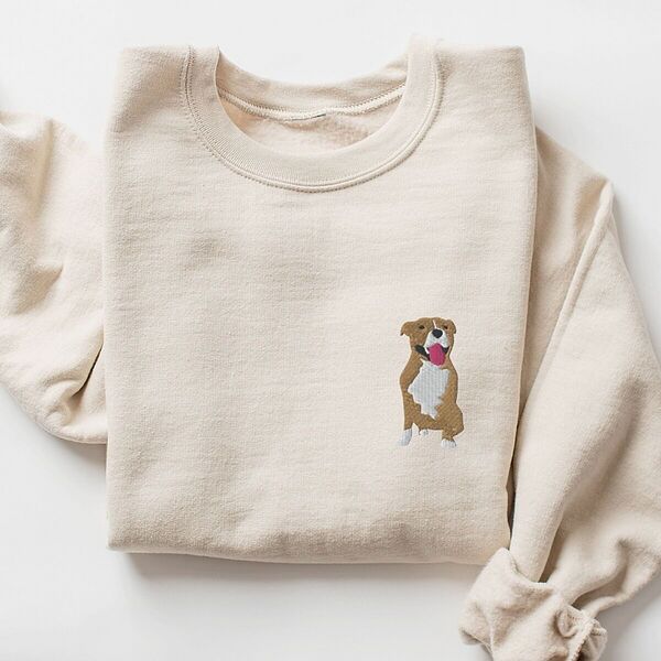 Sand crewneck sweatshirt laid flat with a rolled cuff, embroidered with a small fawn and white pit bull sitting with its pink tongue out on the left chest