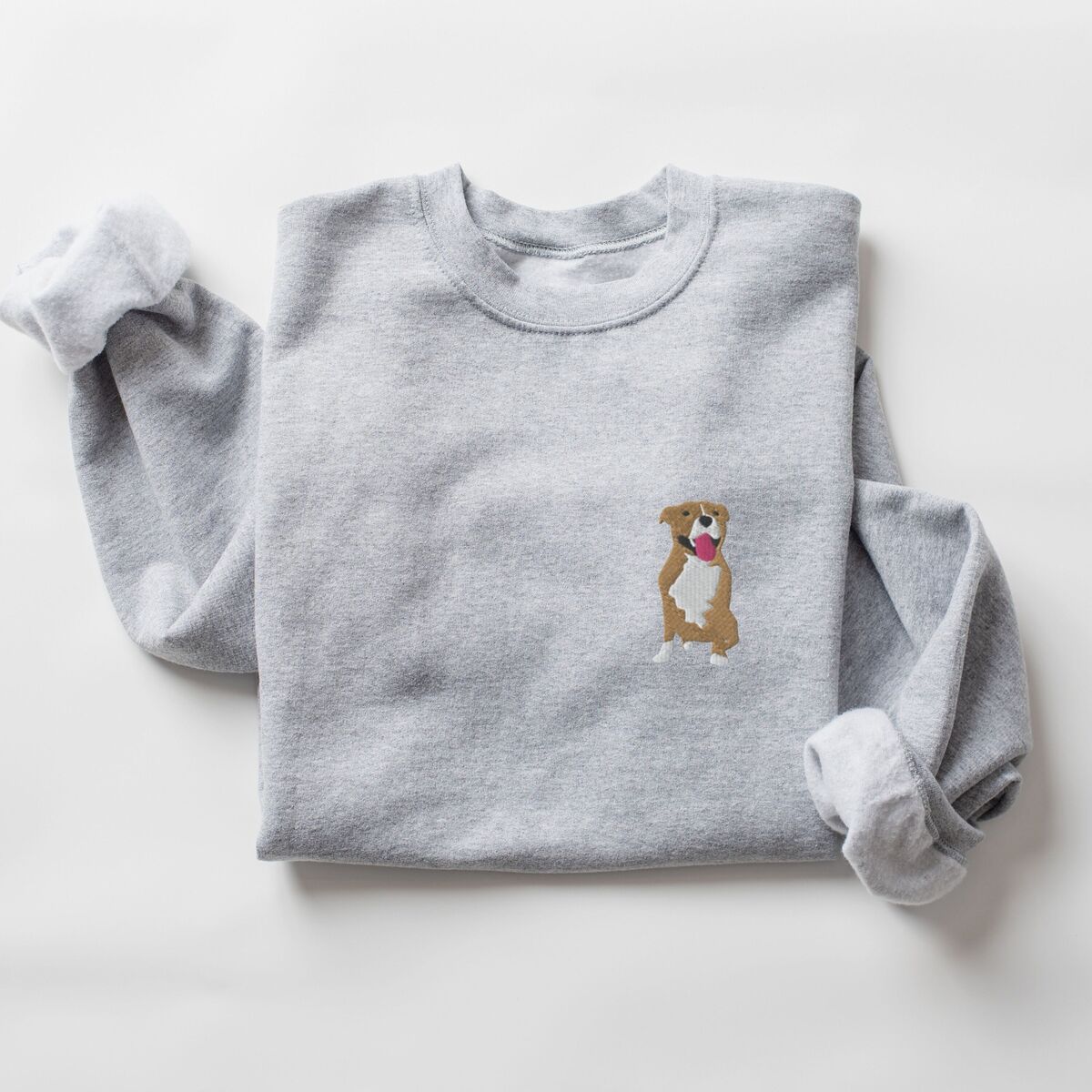 Brown and White Pitbull Sweatshirt - Sport Grey Sport Grey crewneck sweatshirt laid flat with a rolled cuff, embroidered with a small fawn and white pit bull sitting with its pink tongue out on the left chest