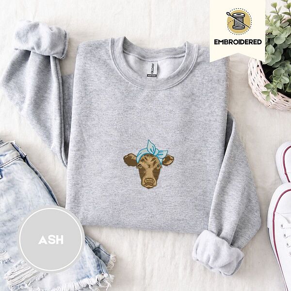Ash crewneck sweatshirt folded with denim shorts, white sneakers and a potted plant, embroidered with a brown calf head wearing a light blue bandana tied in a bow