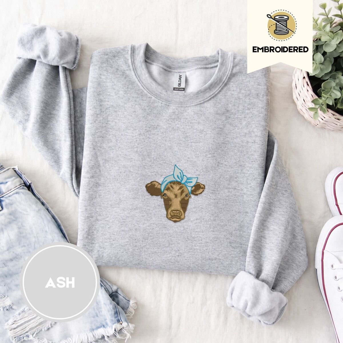 Brown Cow Sweatshirt – Ash Ash crewneck sweatshirt folded with denim shorts, white sneakers and a potted plant, embroidered with a brown calf head wearing a light blue bandana tied in a bow