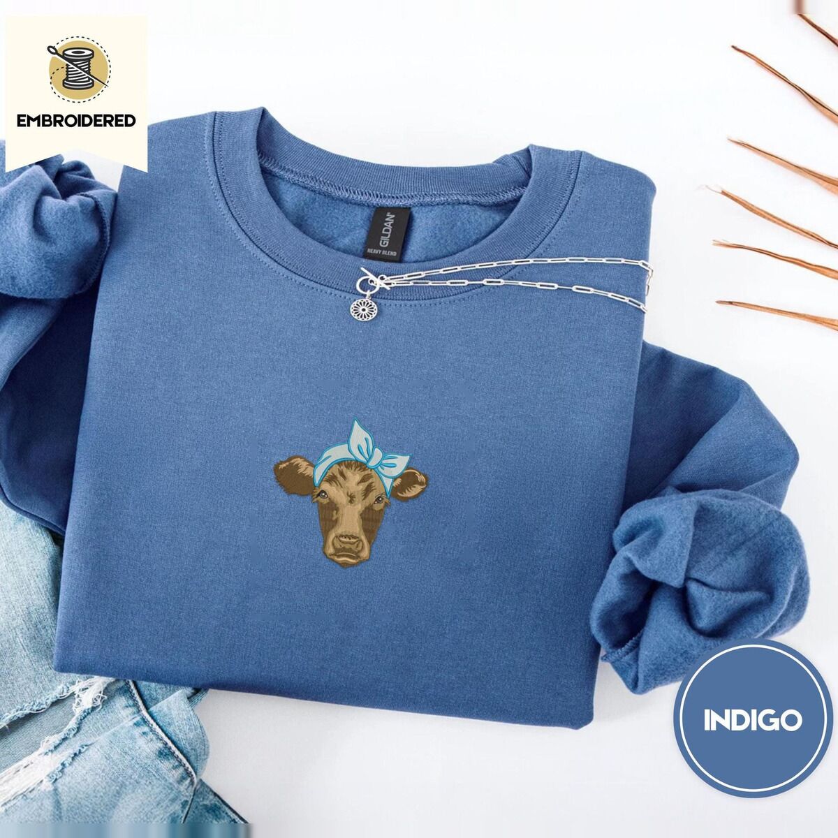 Brown Cow Sweatshirt – Indigo Blue Indigo Blue crewneck sweatshirt folded with light jeans and dried twigs, embroidered with a brown calf head wearing a light blue bandana tied in a bow