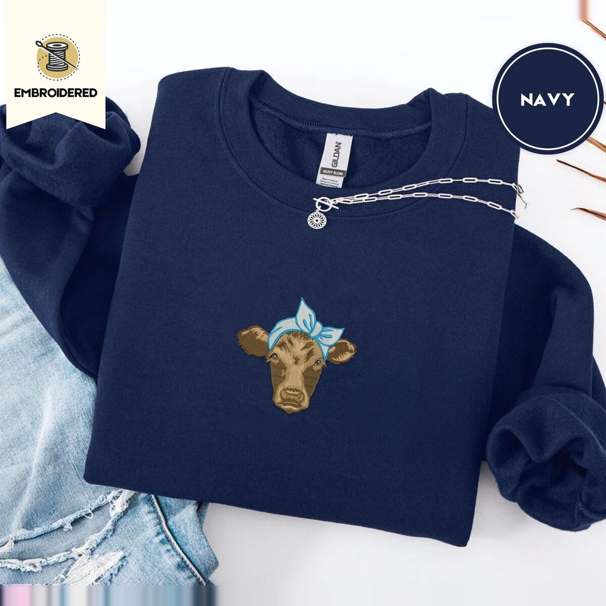 Brown Cow Sweatshirt – Navy Navy crewneck sweatshirt folded with ripped jeans and a silver chain necklace, embroidered with a brown calf head wearing a light blue bandana tied in a bow