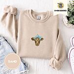 Sand crewneck sweatshirt folded with denim shorts, white sneakers and a potted plant, embroidered with a brown calf head wearing a light blue bandana tied in a bow