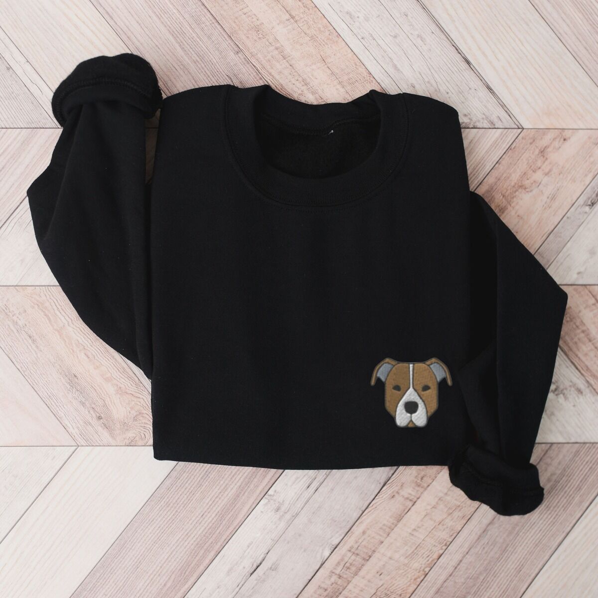 Brown Pitbull Face Sweatshirt – Black Black crewneck sweatshirt laid flat on a wood floor, embroidered with a small minimalist fawn and white pit bull face with grey ears on the left chest