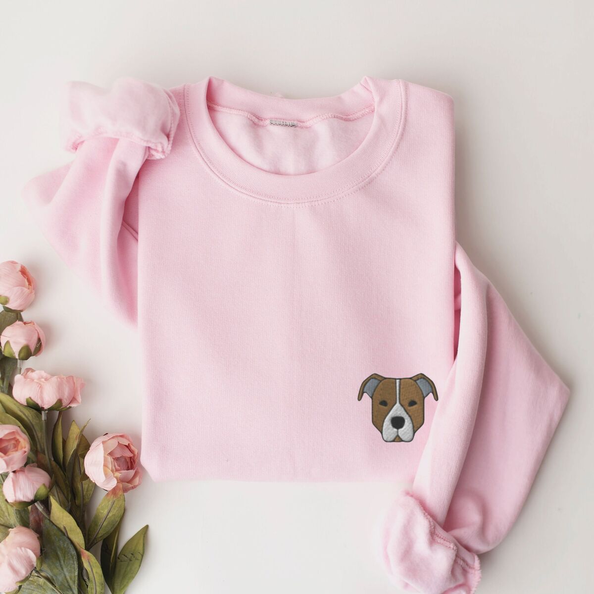 Brown Pitbull Face Sweatshirt - Light Pink Light Pink crewneck sweatshirt laid flat with pink roses, embroidered with a small minimalist fawn and white pit bull face with grey ears on the left chest