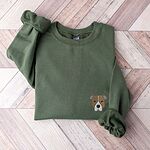 Military Green crewneck sweatshirt laid flat on a wood floor, embroidered with a small minimalist fawn and white pit bull face with grey ears on the left chest