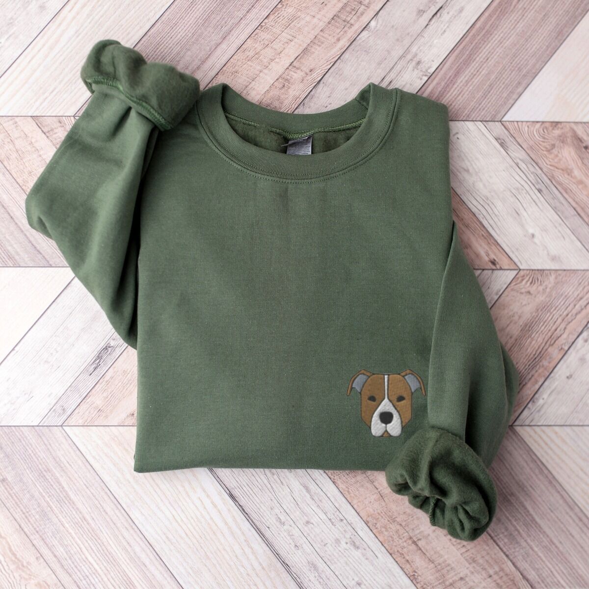 Brown Pitbull Face Sweatshirt – Military Green Military Green crewneck sweatshirt laid flat on a wood floor, embroidered with a small minimalist fawn and white pit bull face with grey ears on the left chest
