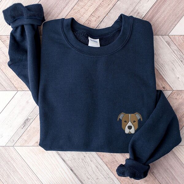 Navy crewneck sweatshirt laid flat on a wood floor, embroidered with a small minimalist fawn and white pit bull face with grey ears on the left chest