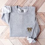 Sport Grey crewneck sweatshirt laid flat on a wood floor, embroidered with a small minimalist fawn and white pit bull face with grey ears on the left chest