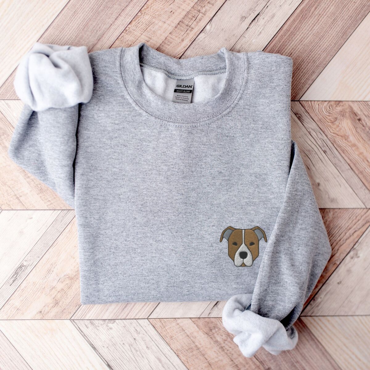 Brown Pitbull Face Sweatshirt - Sport Grey Sport Grey crewneck sweatshirt laid flat on a wood floor, embroidered with a small minimalist fawn and white pit bull face with grey ears on the left chest