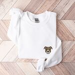 White crewneck sweatshirt laid flat on a wood floor, embroidered with a small minimalist fawn and white pit bull face with grey ears on the left chest