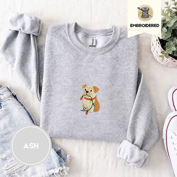 Ash crewneck sweatshirt folded with denim shorts, white sneakers and a potted plant, embroidered with a fawn and cream English bulldog puppy in a red collar chewing a string of Christmas lights
