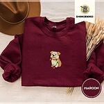 Maroon crewneck sweatshirt with a brown felt hat and dried bunny tail grass, embroidered with a fawn and cream English bulldog puppy in a red collar chewing a string of Christmas lights