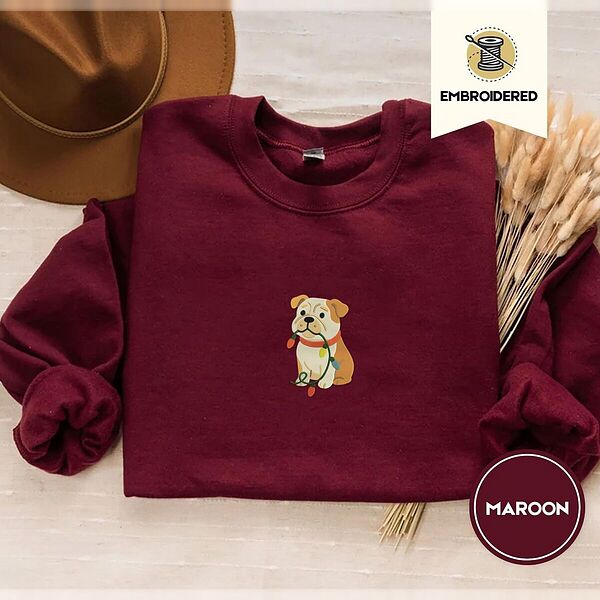 Maroon crewneck sweatshirt with a brown felt hat and dried bunny tail grass, embroidered with a fawn and cream English bulldog puppy in a red collar chewing a string of Christmas lights
