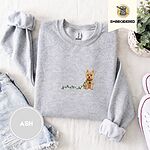 Ash crewneck sweatshirt folded with denim shorts, white sneakers and a potted plant, embroidered with a wheaten Cairn terrier puppy in a red collar chewing the plug of a string of Christmas lights