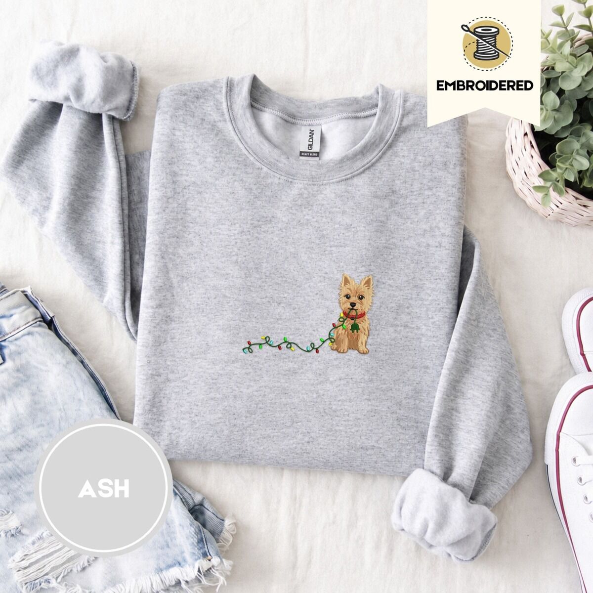 Cairn Terrier Christmas Sweatshirt – Ash Ash crewneck sweatshirt folded with denim shorts, white sneakers and a potted plant, embroidered with a wheaten Cairn terrier puppy in a red collar chewing the plug of a string of Christmas lights