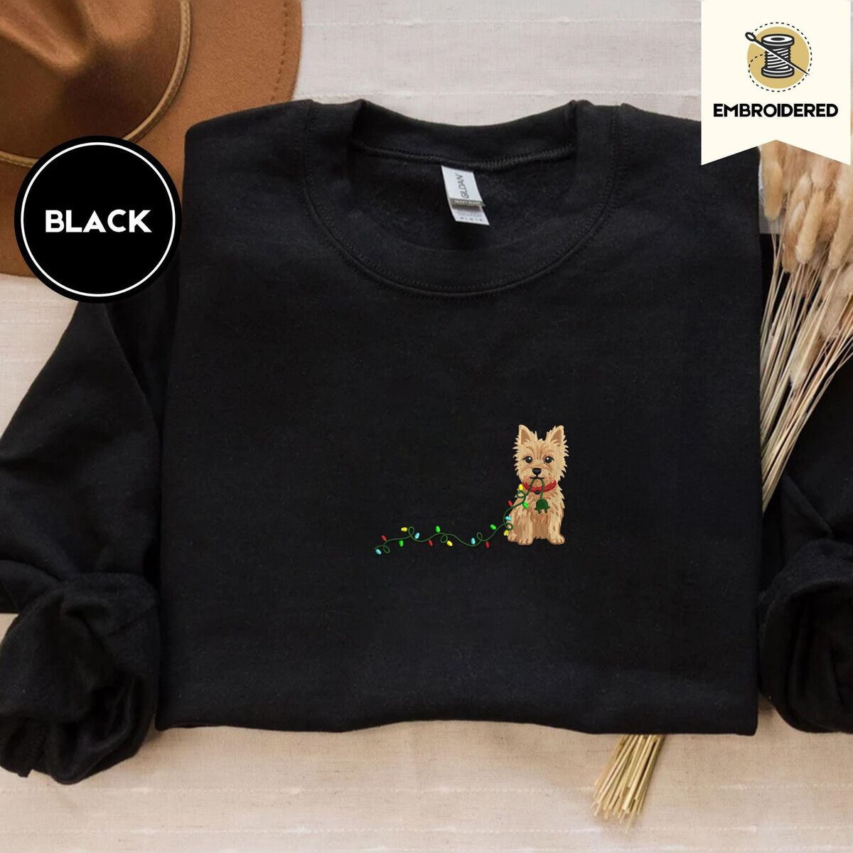 Cairn Terrier Christmas Sweatshirt – Black Black crewneck sweatshirt with a brown felt hat and dried bunny tail grass, embroidered with a wheaten Cairn terrier puppy in a red collar chewing the plug of a string of Christmas lights