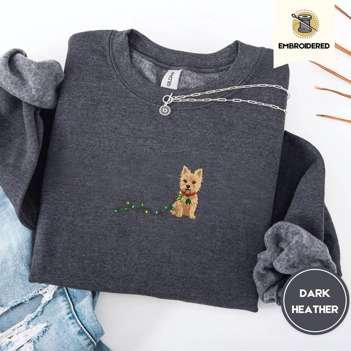 Cairn Terrier Christmas Sweatshirt – Dark Heather Dark Heather crewneck sweatshirt folded with ripped jeans and a silver chain necklace, embroidered with a wheaten Cairn terrier puppy in a red collar chewing the plug of a string of Christmas lights