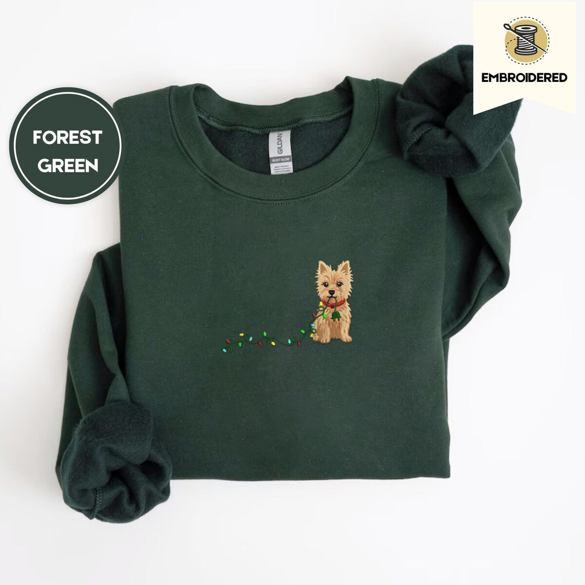 Cairn Terrier Christmas Sweatshirt – Forest Green Forest Green crewneck sweatshirt folded on a white background, embroidered with a wheaten Cairn terrier puppy in a red collar chewing the plug of a string of Christmas lights