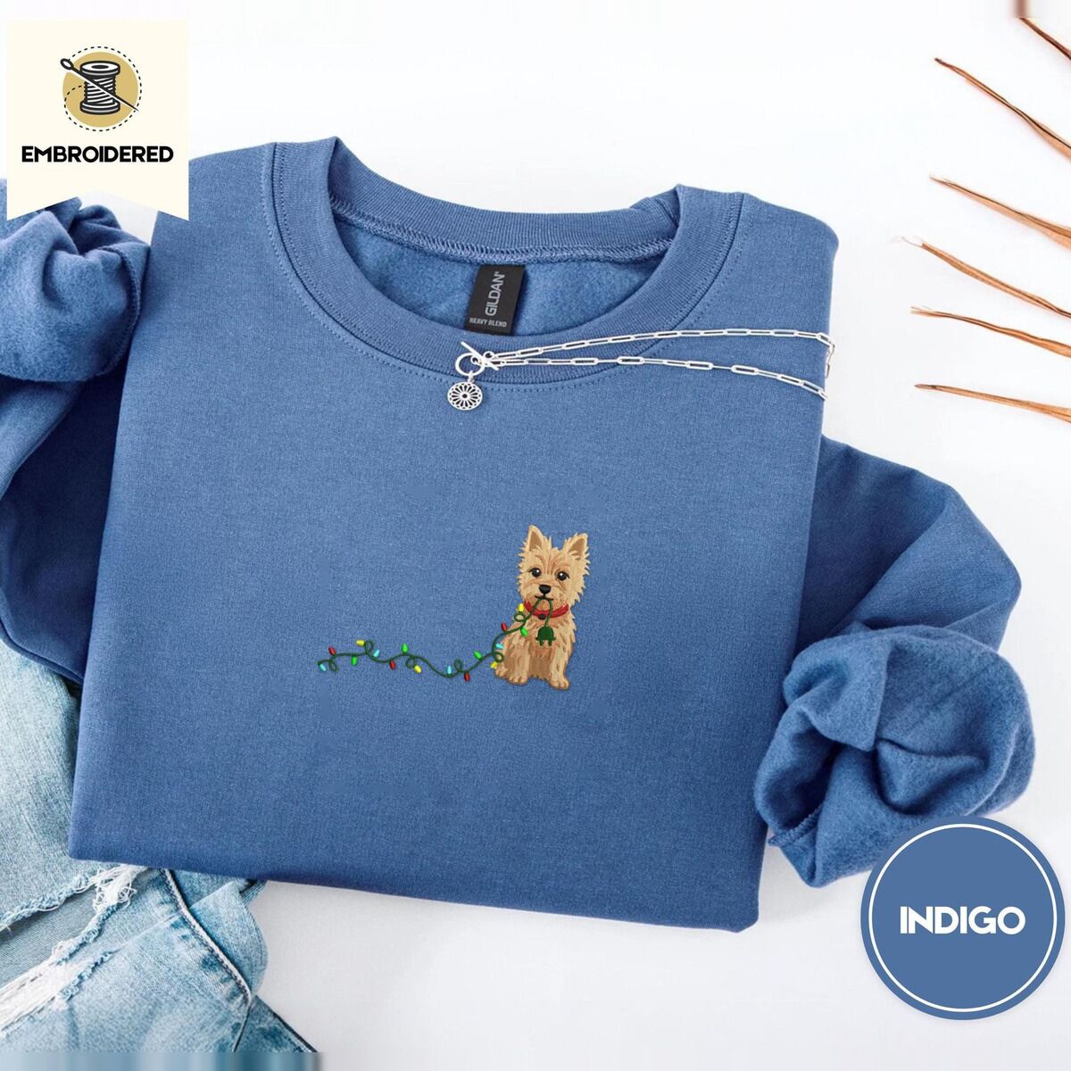 Cairn Terrier Christmas Sweatshirt – Indigo Blue Indigo Blue crewneck sweatshirt folded with light jeans and dried twigs, embroidered with a wheaten Cairn terrier puppy in a red collar chewing the plug of a string of Christmas lights