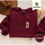 Maroon crewneck sweatshirt with a brown felt hat and dried bunny tail grass, embroidered with a wheaten Cairn terrier puppy in a red collar chewing the plug of a string of Christmas lights
