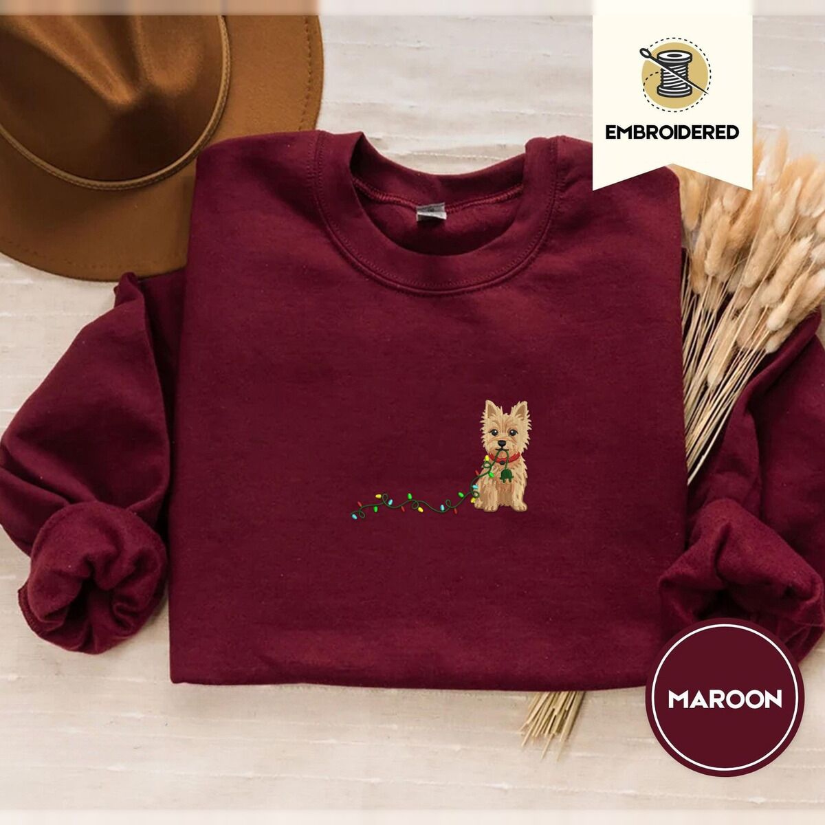 Cairn Terrier Christmas Sweatshirt – Maroon Maroon crewneck sweatshirt with a brown felt hat and dried bunny tail grass, embroidered with a wheaten Cairn terrier puppy in a red collar chewing the plug of a string of Christmas lights