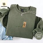 Military Green crewneck sweatshirt folded with light jeans and a silver chain necklace, embroidered with a wheaten Cairn terrier puppy in a red collar chewing the plug of a string of Christmas lights