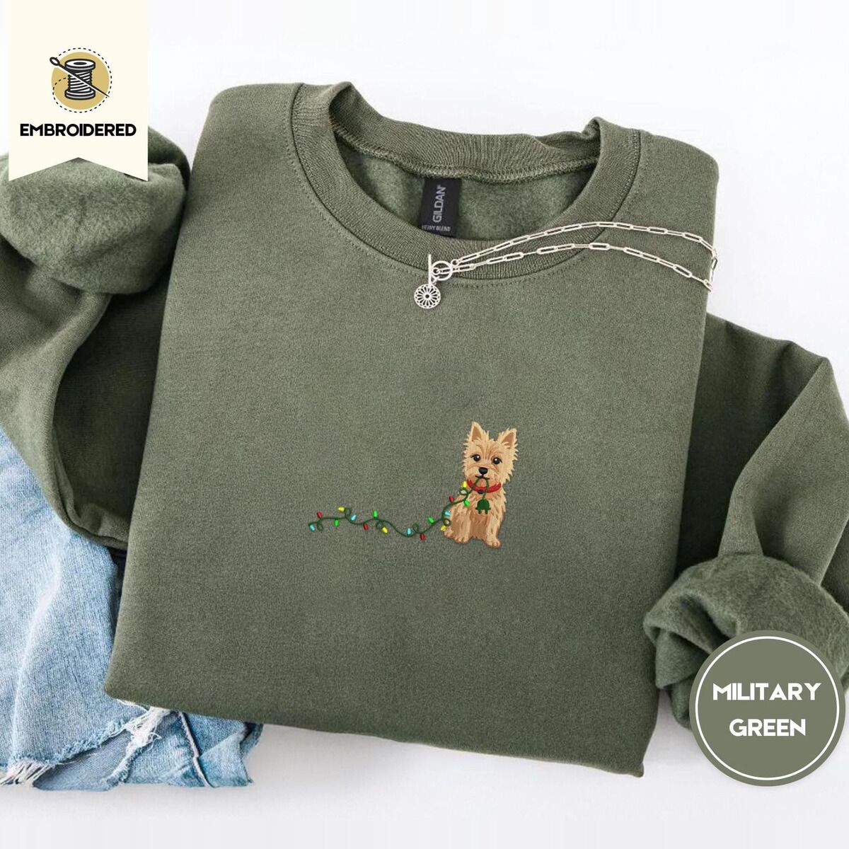 Cairn Terrier Christmas Sweatshirt – Military Green Military Green crewneck sweatshirt folded with light jeans and a silver chain necklace, embroidered with a wheaten Cairn terrier puppy in a red collar chewing the plug of a string of Christmas lights