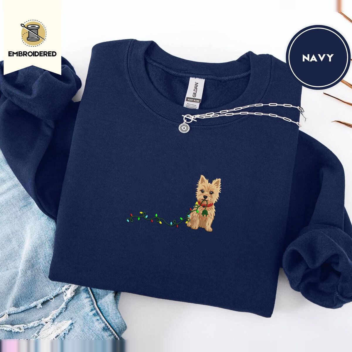 Cairn Terrier Christmas Sweatshirt – Navy Navy crewneck sweatshirt folded with ripped jeans and a silver chain necklace, embroidered with a wheaten Cairn terrier puppy in a red collar chewing the plug of a string of Christmas lights