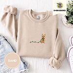 Sand crewneck sweatshirt folded with denim shorts, white sneakers and a potted plant, embroidered with a wheaten Cairn terrier puppy in a red collar chewing the plug of a string of Christmas lights