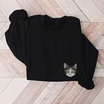 Black crewneck sweatshirt folded on a herringbone wood floor, with a calico cat face with green eyes embroidered on the left chest