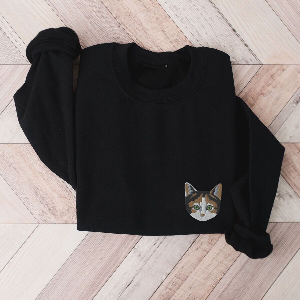 Calico Cat Sweatshirt – Black Black crewneck sweatshirt folded on a herringbone wood floor, with a calico cat face with green eyes embroidered on the left chest