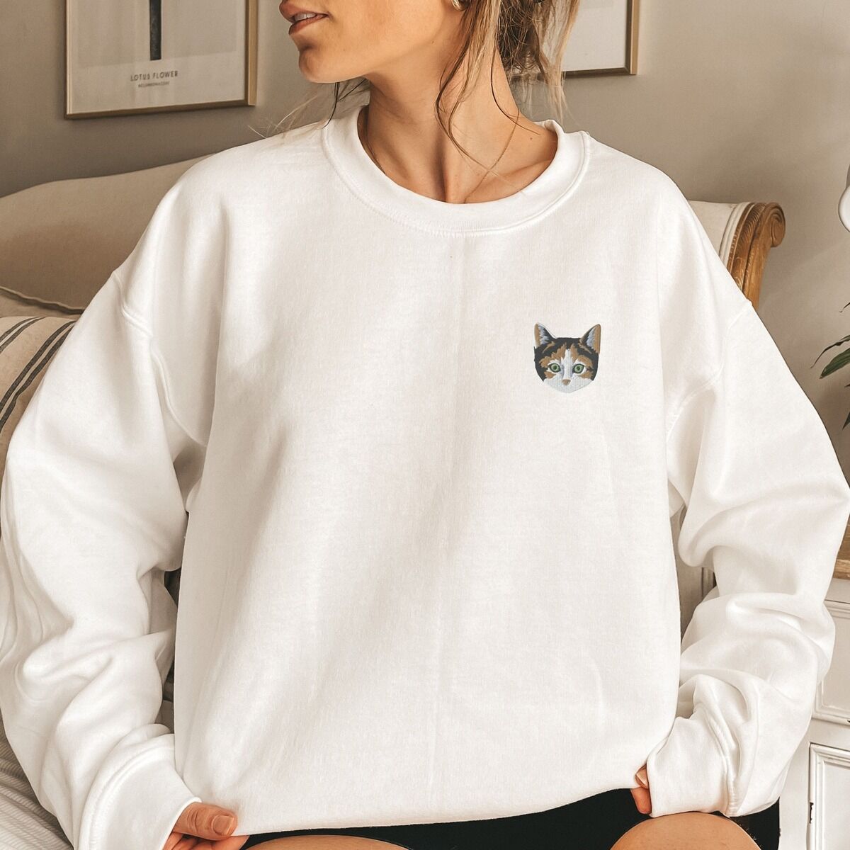 Calico Cat Sweatshirt Worn White Woman sitting on a couch in a White crewneck sweatshirt, with a calico cat face with green eyes embroidered on the left chest