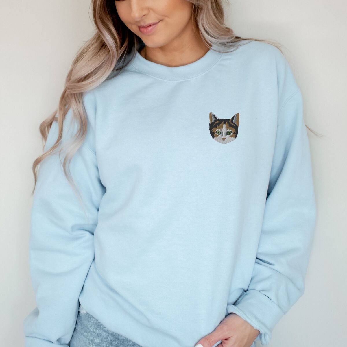 Calico Cat Sweatshirt Light Blue Woman with long wavy hair in a Light Blue crewneck sweatshirt, with a calico cat face with green eyes embroidered on the left chest